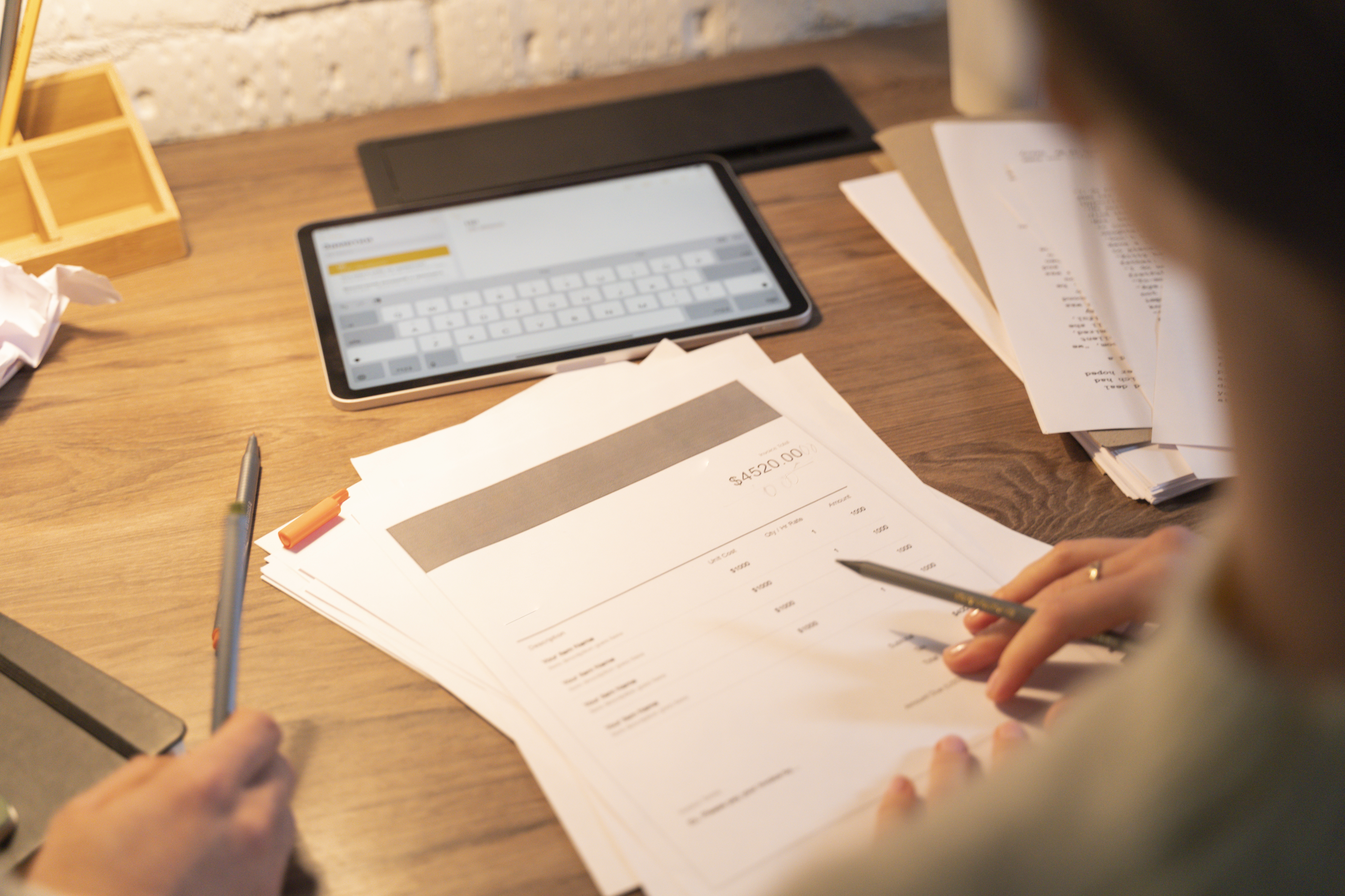 Overhead view of a person manually processing paper invoices at a desk with a tablet, highlighting the need for invoice scanning software.