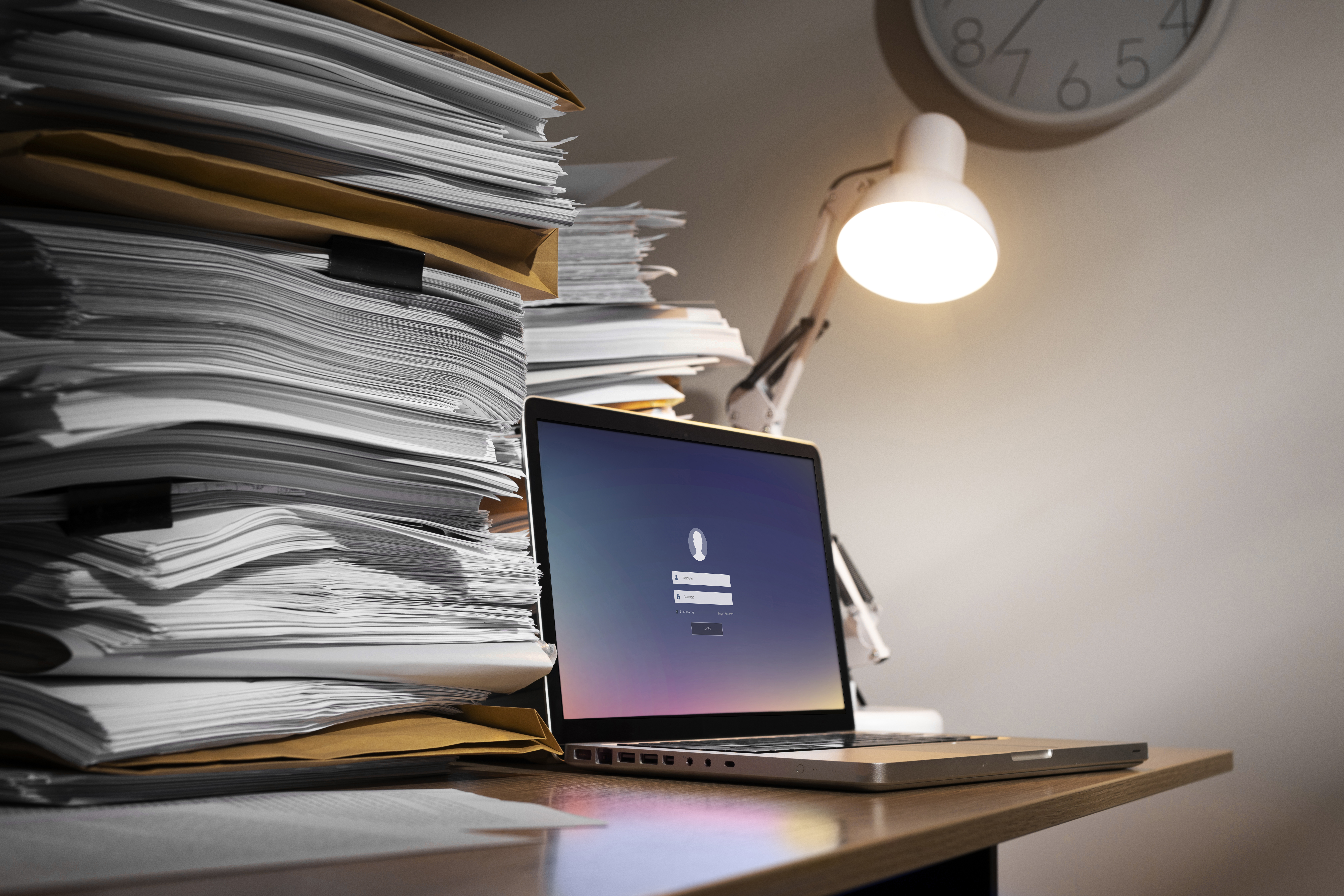 A laptop displaying a login screen sits beside a tall stack of paper documents, representing the shift from manual to digital invoice reconciliation.
